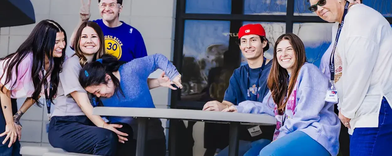 Staff and members laugh and goof around together at an outdoor table, capturing friendship and joy in a relaxed day program setting.